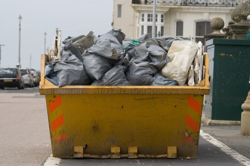 A large yellow skip situated on an urban street pavement, filled with black and white rubbish bags made of plastic and fabric, some tightly packed and others slightly bulging. The skip is positioned near a white building with windows and decorative elements, with a green trash bin visible on the right side of the image. In the background, parked cars and overcast sky suggest a typical street scene in Willesden, London. The skip displays signs of dirt and minor wear, indicating recent use for rubbish removal, aligning with services provided by Willesden House Clearance in the area. The scene captures a practical aspect of waste collection in a residential or commercial setting, emphasizing the importance of efficient rubbish disposal in the local community.