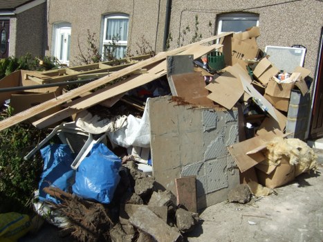 A pile of mixed household rubbish and building debris situated on a pavement in front of a residential building, involving various materials such as cardboard boxes, wooden planks, concrete blocks, and fabric coverings. The cardboard boxes are flattened and scattered, with some partially crushed or torn, in shades of brown and beige. Wooden planks, some with rough textures, lean against the pile, showing signs of splintering and weathering, with light wood tones. Concrete blocks with a rough, grey surface are stacked irregularly, intermingled with smaller debris. Blue plastic bags and white fabric coverings are visible, adding colour variation and indicating different types of waste. To the left, some machinery or tools are partially visible, possibly related to rubbish collection. The background shows the exterior wall and windows of a residential property, with a small garden area partly visible. Overall, the scene captures a typical rubbish accumulation for clearance services like those offered by Willesden House Clearance, emphasizing the need for professional waste removal in the local area.