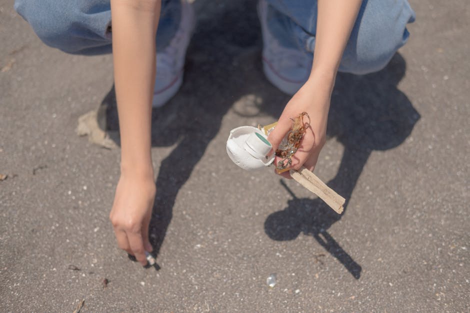 A person wearing light blue jeans and white sneakers is crouching on a paved surface outdoors, holding a small makeshift model of a house made from a white plastic cup, a piece of wood, and various sc