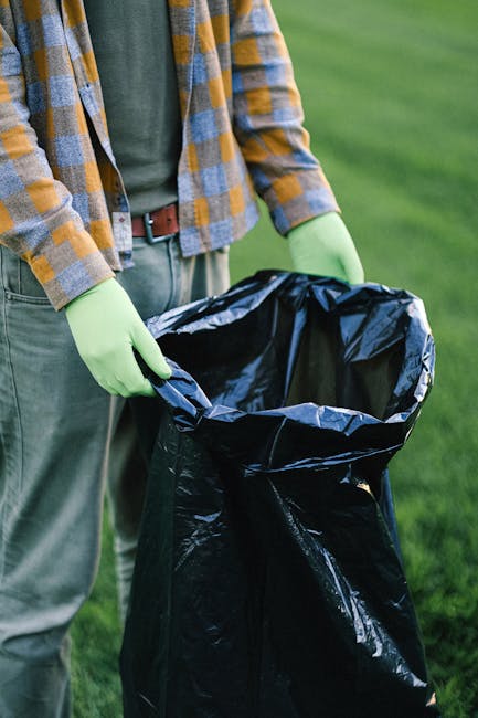 A person wearing light blue jeans and white sneakers is crouching on a paved surface outdoors, holding a small makeshift model of a house made from a white plastic cup, a piece of wood, and various sc