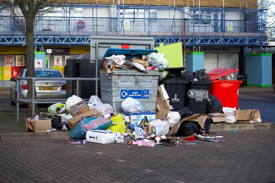Two large black plastic bags filled with waste are placed on the pavement beside a black metal fence with vertical bars. The bags appear to contain bulky rubbish, possibly including paper or mixed mat
